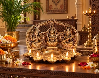 Puede incluir: Un centro de mesa decorativo de metal dorado con tres deidades sentadas, Ganesha, Lakshmi y Saraswati, dispuestas en fila. La base incluye cinco portavelas integrados con llamas encendidas y un cuenco circular central para ofrendas.