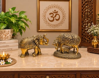 Puede incluir: Una figura de elefante en tono dorado metálico con patrones en relieve intrincados y una figura de vaca con ternero en tono dorado metálico sobre una base ovalada con tallas florales detalladas, ambas sobre una superficie clara con una vela encendida.