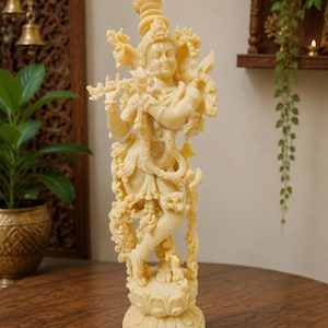 May include: A cream-coloured statue of a deity standing on a lotus base, playing a flute. The figure is adorned with intricate jewellery, a crown, and flowing garments, with a peacock perched near the shoulder, all rendered in a smooth, carved finish.