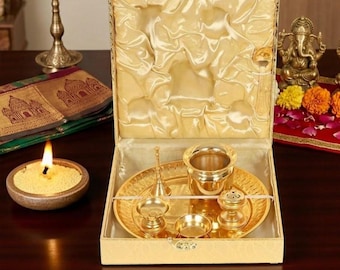 Puede incluir: Un juego de puja thali de metal dorado presentado en una caja forrada de tela color crema. El conjunto incluye una bandeja circular, una taza pequeña, una campana y dos cuencos pequeños. El interior de la caja tiene tela satinada fruncida.