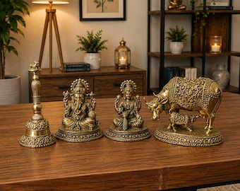 Puede incluir: Un conjunto de cuatro objetos decorativos de color latón que incluye una campana alta ornamentada, una figura de Ganesha sentada, una figura de Lakshmi sentada y una figura de vaca con ternero, con detalles en relieve.