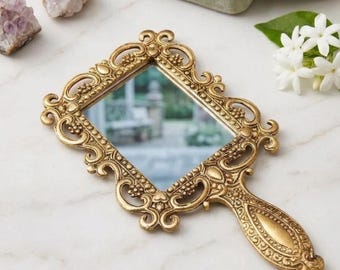 Puede incluir: Un espejo de mano rectangular con un marco dorado ornamentado que presenta intrincados trabajos de volutas y detalles de cuentas. El mango es cónico con un panel central liso y bordes decorativos. La superficie reflectante muestra una escena exterior borrosa.