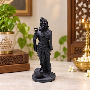 Könnte beinhalten: Eine mattschwarze Statue einer Gottheit auf einem runden Sockel, die eine goldfarbene Flöte spielt. Die Figur trägt traditionelle drapierte Kleidung, Halsketten und einen kleinen roten Punkt auf der Stirn. Ein kleiner, strukturierter Topf steht zu Füßen.