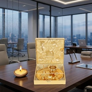 May include: A gold-colored metal set consisting of a round tray, a small bowl, a bell, and a small container, all housed in a square, open-top box with a gold satin-lined interior. A small, round, beige candle in a wooden bowl sits nearby.