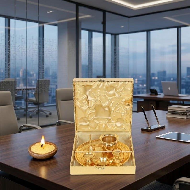 May include: A gold-colored metal set consisting of a round tray, a small bowl, a bell, and a small container, all housed in a square, open-top box with a gold satin-lined interior. A small, round, beige candle in a wooden bowl sits nearby.