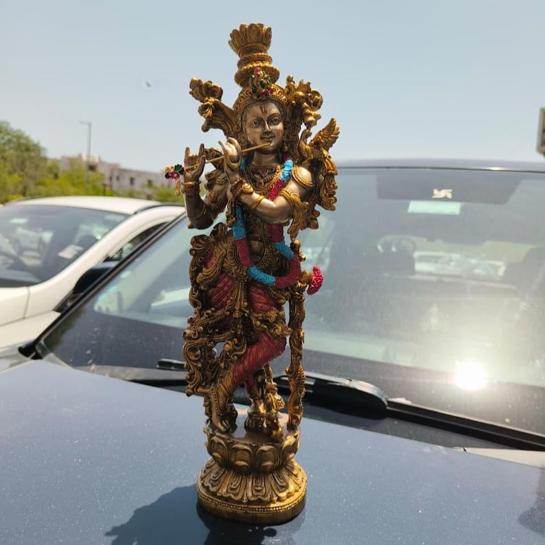 Puede incluir: Una estatua dorada detallada de una deidad tocando la flauta. La estatua presenta intrincadas tallas, detalles en rojo y azul, y est&aacute; montada sobre una base decorativa. La figura est&aacute; adornada con joyas y una corona.
