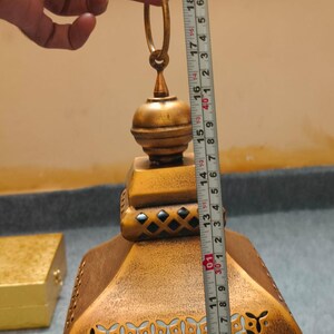Puede incluir: Una linterna de metal dorado con un patr&oacute;n decorativo recortado y un anillo superior para colgar. Una cinta m&eacute;trica colocada verticalmente contra la linterna indica una altura de aproximadamente 48 cm desde la base hasta la parte superior del anillo.