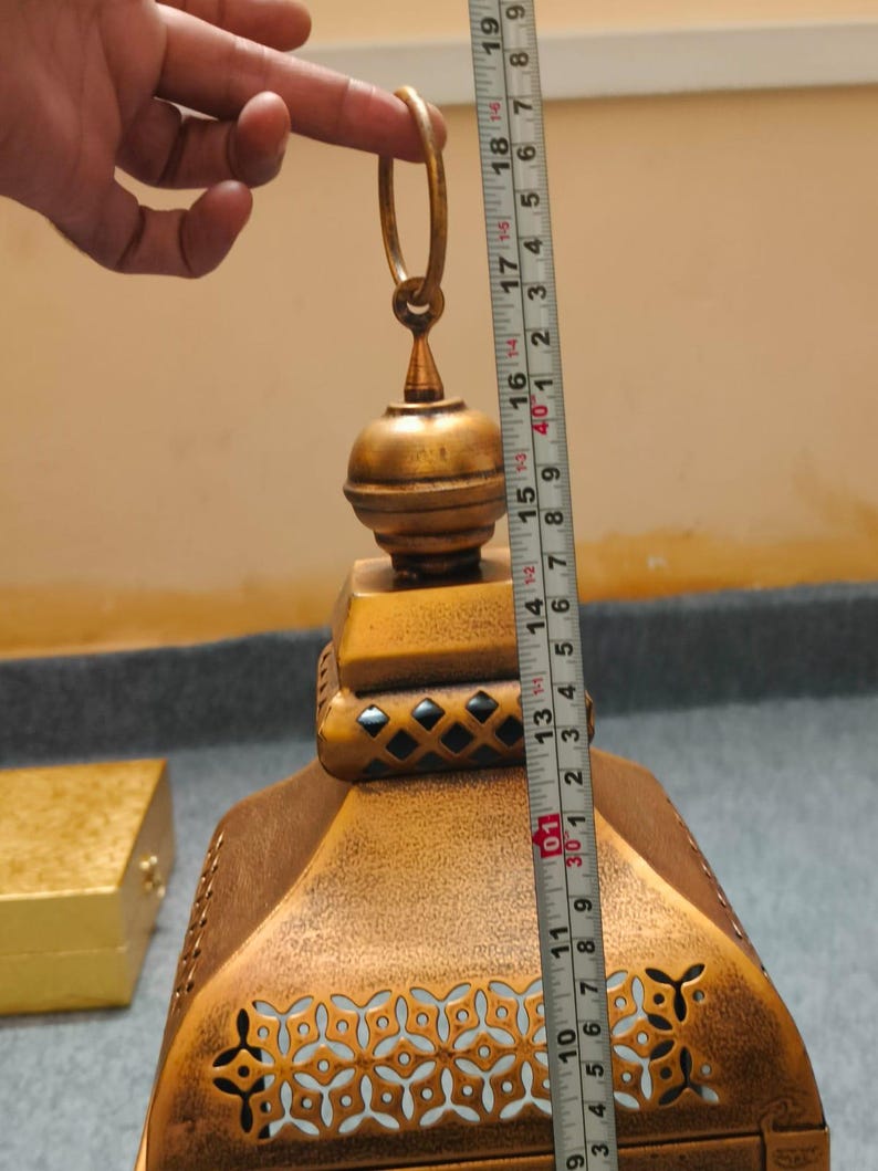 Puede incluir: Una linterna de metal dorado con un patr&oacute;n decorativo recortado y un anillo superior para colgar. Una cinta m&eacute;trica colocada verticalmente contra la linterna indica una altura de aproximadamente 48 cm desde la base hasta la parte superior del anillo.