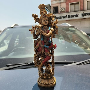 Puede incluir: Una estatua detallada de color bronce de una deidad tocando una flauta. La figura lleva ropa ornamentada en rojo, azul y dorado, con tocados y joyas elaboradas. La estatua est&aacute; sobre una base decorativa, que muestra intrincadas tallas.