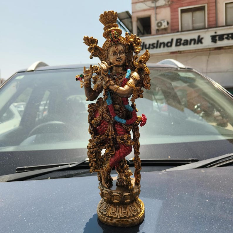 Puede incluir: Una estatua detallada de color bronce de una deidad tocando una flauta. La figura lleva ropa ornamentada en rojo, azul y dorado, con tocados y joyas elaboradas. La estatua est&aacute; sobre una base decorativa, que muestra intrincadas tallas.