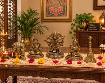 Puede incluir: Una colección de cinco objetos decorativos de color latón que incluye una figura de elefante, tres estatuas de deidades y una campana ritual. Los artículos presentan intrincados patrones en relieve y están dispuestos sobre un camino de mesa con flores.