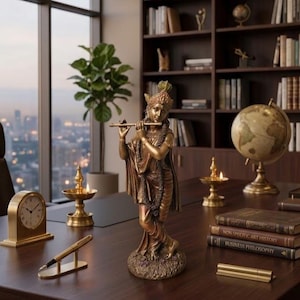 May include: A bronze-toned statue of a figure standing on a base, playing a flute. The figure wears an ornate headdress and draped clothing. The statue is positioned on a dark wooden desk alongside a clock, a pen, two lit oil lamps, and books.