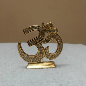 Könnte beinhalten: Eine goldfarbene Om-Symbol-Figur mit poliertem Metallfinish, die mit zahlreichen kleinen, klaren und funkelnden Strasssteinen besetzt ist. Das Dekorationsobjekt steht auf einem rechteckigen Sockel und weist eine glatte, reflektierende Oberfläche auf.