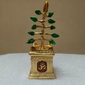 Könnte beinhalten: Eine goldfarbene Metallskulptur eines Baumes mit grün bemalten Blättern, die auf einem quadratischen Sockel mit einem geprägten roten Om-Symbol steht. Der Sockel weist auf allen Seiten filigrane Verzierungen auf und der Baum hat eine goldene Spitze.
