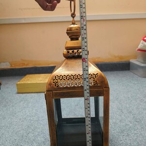 Puede incluir: Un farol de metal dorado con paneles de vidrio y patrones recortados intrincados cerca de la parte superior. Una cinta m&eacute;trica colocada contra el farol muestra una altura total de aproximadamente 48 cm desde la base hasta la parte superior del anillo.