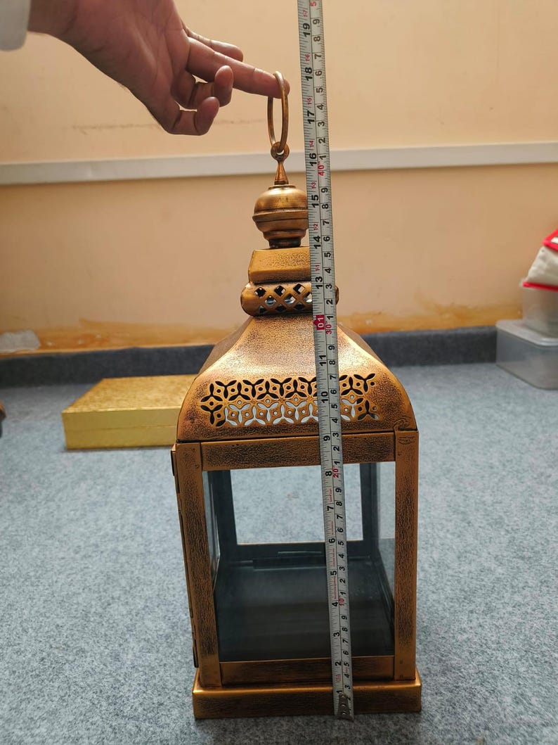 Puede incluir: Un farol de metal dorado con paneles de vidrio y patrones recortados intrincados cerca de la parte superior. Una cinta m&eacute;trica colocada contra el farol muestra una altura total de aproximadamente 48 cm desde la base hasta la parte superior del anillo.