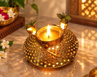 Puede incluir: Un portavelas de metal dorado con una base en forma de cúpula que presenta un patrón de recortes repetitivos. El centro superior sostiene una vela encendida, flanqueada por dos elementos decorativos de metal con forma de pequeños frutos.