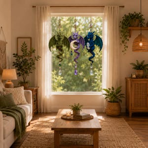 May include: Three hanging dragon ornaments suspended in a window. The left dragon is green, the center is purple holding a white crescent moon, and the right is blue holding a clear crystal. Each features a faceted glass prism at the bottom.