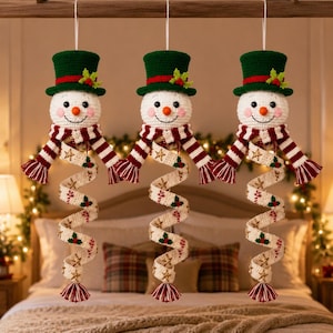 Peut inclure: Trois décorations de bonhomme de neige au crochet suspendues à une barre en bois. Chaque bonhomme porte un haut-de-forme vert avec une bande rouge et du houx, une tête blanche avec un nez carotte et une écharpe rayée rouge et blanche.