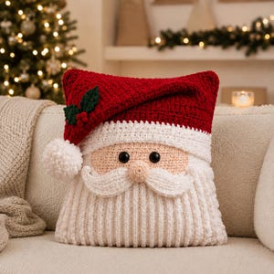 May include: A square crochet pillow shaped like a Santa Claus face. The top features a red hat with a white pom-pom and green holly leaves with red berries. The face has a peach-colored skin tone, black button eyes, and a white textured beard.