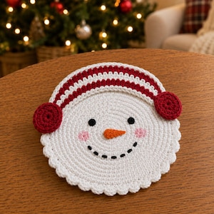 May include: A round, white crocheted snowman face coaster featuring a red and white striped earmuff headband, two black circular eyes, a small orange carrot-shaped nose, a black stitched smile, and two light pink circular blush marks.