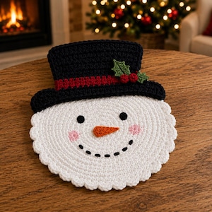 May include: A round, white crocheted snowman face with a scalloped edge, featuring two black circular eyes, a stitched black smile, pink cheeks, and an orange nose. It wears a black crocheted top hat with a red band and green holly leaves.