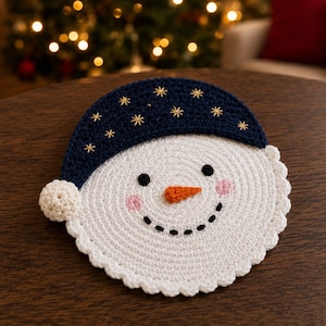 May include: A circular crochet snowman face coaster featuring a white base with a scalloped edge, a dark blue hat with embroidered gold star accents, a small white pom-pom, black circular eyes, a stitched black smile, and an orange nose.