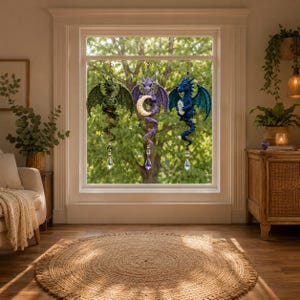 May include: Three crocheted dragon ornaments hanging in a window. The left dragon is green, the center is purple holding a crescent moon, and the right is blue holding a clear crystal. Each dragon features a dangling faceted crystal prism.