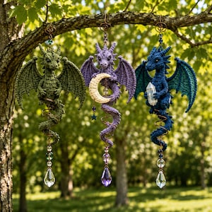 May include: Three crocheted dragon ornaments hanging from a tree branch. The left is olive green, the center is purple holding a crescent moon, and the right is blue holding a clear crystal. Each features a spiral wire tail and a crystal prism.