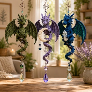 May include: Three hanging crochet dragon ornaments in olive green, purple, and dark blue. Each dragon features textured wings, a coiled tail, and a dangling crystal prism. The purple dragon holds a crescent moon, while the blue one holds a crystal.