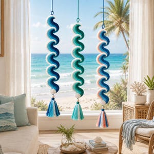 Può includere: Tre decorazioni verticali da appendere con un motivo a onda a spirale realizzato in filato. Ogni pezzo presenta una sfumatura di colori blu, verde acqua o pesca, rifinito con una conchiglia bianca e una lunga nappa in fibra multicolore.