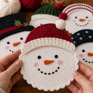 May include: A round, white crocheted snowman coaster featuring a red hat with a white pom-pom, black stitched eyes, a small orange carrot nose, and pink cheeks. The edge of the coaster has a scalloped design, and it is held against a background of other snowman-themed crochet coasters.