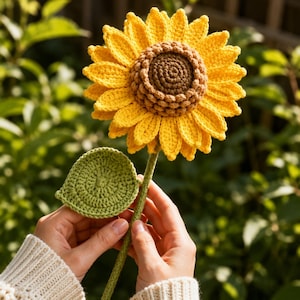 May include: A handcrafted crochet sunflower featuring a textured brown center surrounded by bright yellow petals. The flower is attached to a long green stem with a single round green leaf extending from the side, showcasing detailed yarn stitching.