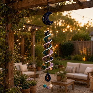 May include: A hanging crochet mobile featuring a dark blue crescent moon at the top with yellow star embroidery. Below, a spiral structure transitions from teal to purple, accented with small yellow stars. A dark blue teardrop shape hangs at the base.