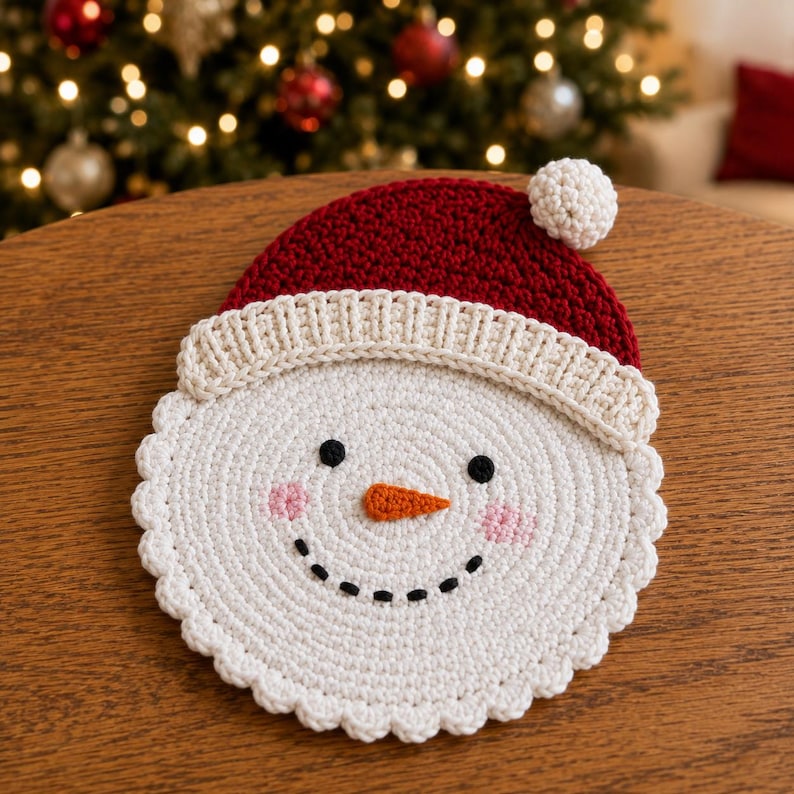 May include: A circular crochet snowman face with a red hat featuring a white pom-pom and brim. The snowman has a white base, two black eyes, a stitched orange carrot nose, a black stitched smile, and pink embroidered cheeks on the sides.