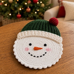 May include: A circular crochet snowman face with a white base, a dark green ribbed hat featuring a red pom-pom, two black eyes, a stitched orange carrot nose, a dotted black smile, and two light pink circular cheeks on the white surface.