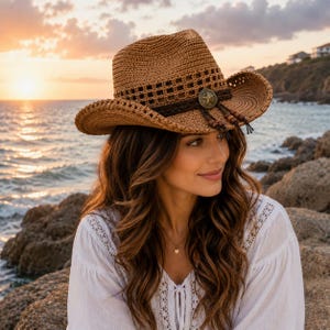 May include: A tan woven straw cowboy hat featuring a dark brown braided band, a circular metal star emblem at the center, and dangling wooden beads. The hat has a structured crown and a wide, slightly curved brim with a decorative open-weave pattern.