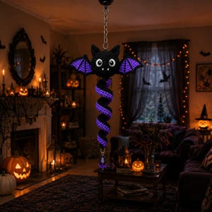 May include: A hanging crochet decoration featuring a black bat with purple wing accents and large white eyes. A long, spiraling black and purple tail extends downward from the bat, ending with a faceted purple crystal pendant attached by a metal chain.
