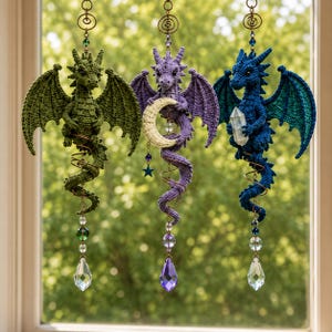 May include: Three hanging crochet dragon ornaments in green, purple, and blue. The purple dragon holds a crescent moon, while the blue dragon holds a clear crystal. Each dragon is suspended by a metal spiral and features a dangling crystal prism.