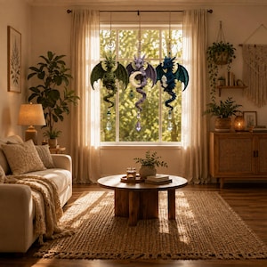 May include: Three hanging dragon ornaments suspended in a window. The left dragon is green, the center is purple holding a crescent moon, and the right is blue holding a crystal. Each dragon features a dangling faceted crystal prism at the base.