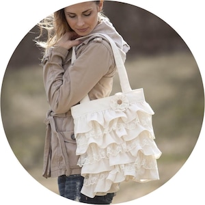 Op de afbeelding: Een witte canvas tote bag met een gerimpelde rand en een bloemversiering. De tas heeft een bruine knoop en is gemaakt van een zachte, lichte stof.