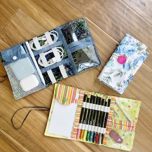 Può includere: Tre organizer in tessuto: una custodia aperta in denim blu con rete bianca ed elastici per cavi, una custodia aperta gialla con bordi a righe per matite colorate e gomma, e una custodia chiusa a fiori con un bottone rosa.