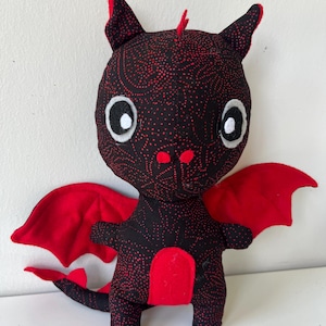 Può includere: Un giocattolo a forma di drago di peluche fatto a mano. Il drago è principalmente nero con un motivo a pois rossi e dettagli rossi. Ha grandi occhi bianchi, ali rosse e una pancia rossa. Il drago è in piedi.