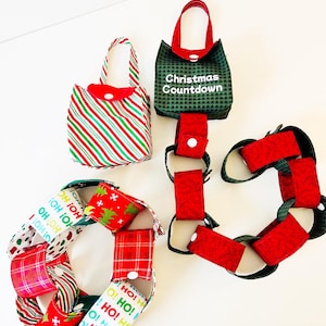 Op de afbeelding: Een kerst-countdown adventkalenderketting gemaakt van stof met rode, groene en witte patronen. De ketting is gemaakt van individuele stoffen zakjes die aan elkaar zijn verbonden. Elk zakje heeft een knoopsluiting. De ketting is versierd met de woorden "Ho! Ho!" en "Christmas Countdown".
