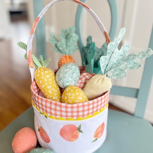 Peut inclure: Un panier de Pâques en tissu blanc avec un imprimé pêche et une bordure en vichy. Le panier est rempli de carottes en tissu, d'œufs et d'une tige en tissu vert feuillage.