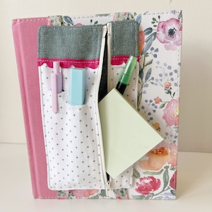 May include: A white and black patterned fabric zipper pouch with a pink trim. The pouch is filled with a green pen, two blue and pink pens, and a light green sticky note.