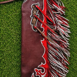 May include: A dark brown leather chaps piece featuring intricate floral tooling at the top, white leather trim, and a vibrant red leather panel. The side is adorned with long, layered fringe strips in alternating shades of red, white, and brown.