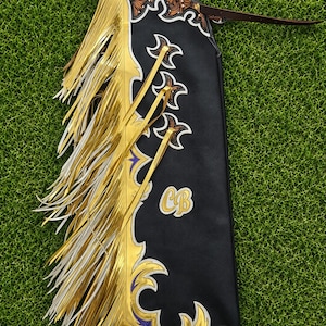 Pode incluir: Chaps de couro preto com longas franjas douradas metalizadas, acabamentos decorativos em branco e roxo e um intrincado trabalho floral gravado no topo. O painel lateral exibe as letras CB bordadas em ouro com detalhes dourados e brancos.