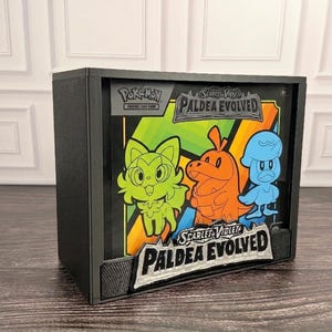 May include: A black display case containing a Pokémon Scarlet and Violet Paldea Evolved trading card game booster box. The front features colorful illustrations of Sprigatito, Fuecoco, and Quaxly, with the text Paldea Evolved clearly visible.