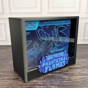 May include: A black display case containing a Pokémon Elite Trainer Box with a blue and black graphic of a creature. The front features a blue flame-shaped sign with the text Mega Evolution Phantasmal Flames in a bold, stylized font.